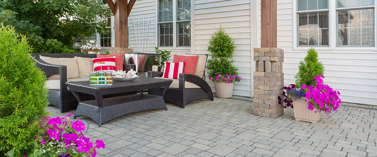Cozy outdoor paver patio with wicker seating under a wooden gazebo featuring decorative pillows and vibrant potted plants.