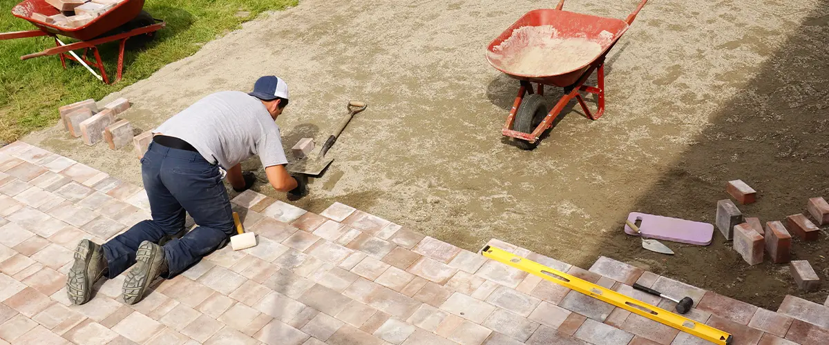 Worker installing paving stones for a new outdoor paver patio with tools and materials including a wheelbarrow and level nearby.