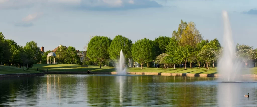 A wide landscape shot of a large community pond featuring three high-pressure floating fountains used for water aeration and decoration.