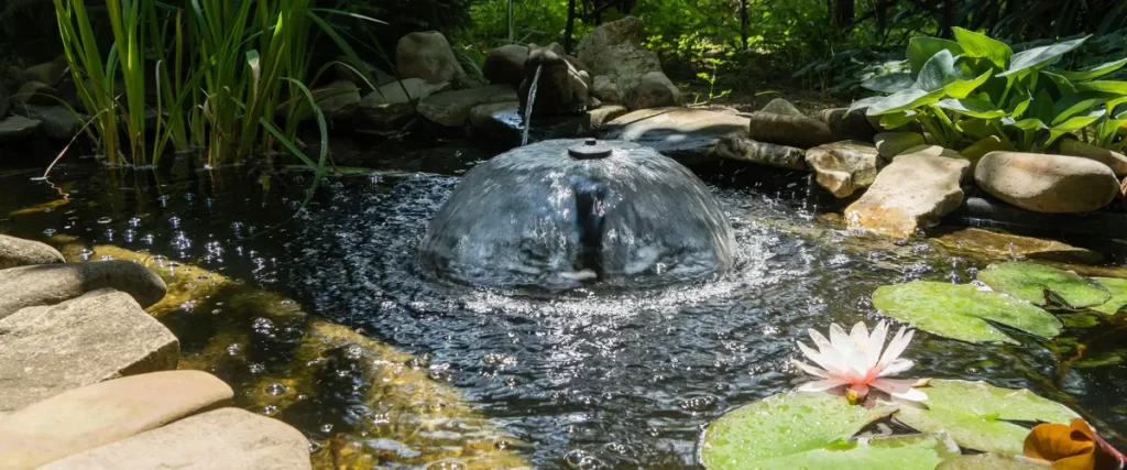 A close-up view of a circular water aerator creating a dome-shaped ripple effect in a stone-lined pond next to a white lotus flower.