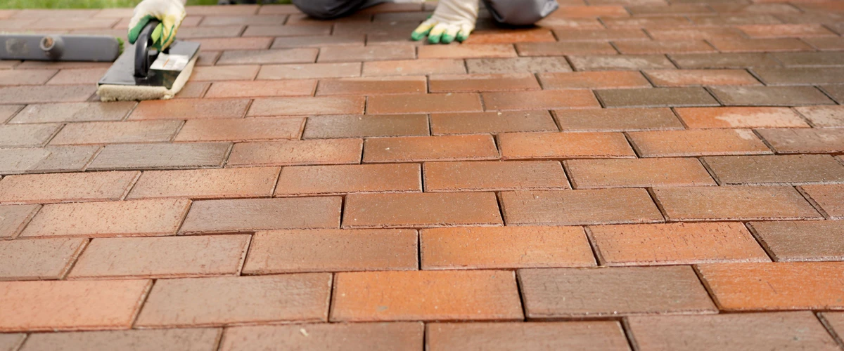 A close-up of a contractor using a hand-held sponge float tool to apply a wet, glossy protective sealant to a multi-colored red and orange brick paver walkway.