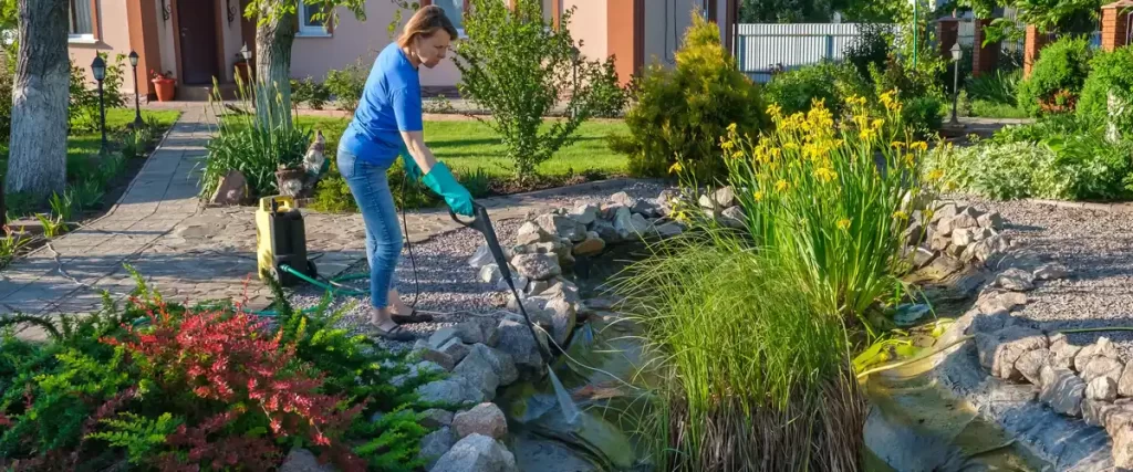 A woman in blue performing koi pond maintenance by using a pressure washer to clean the stone border and liner of an empty garden pond.