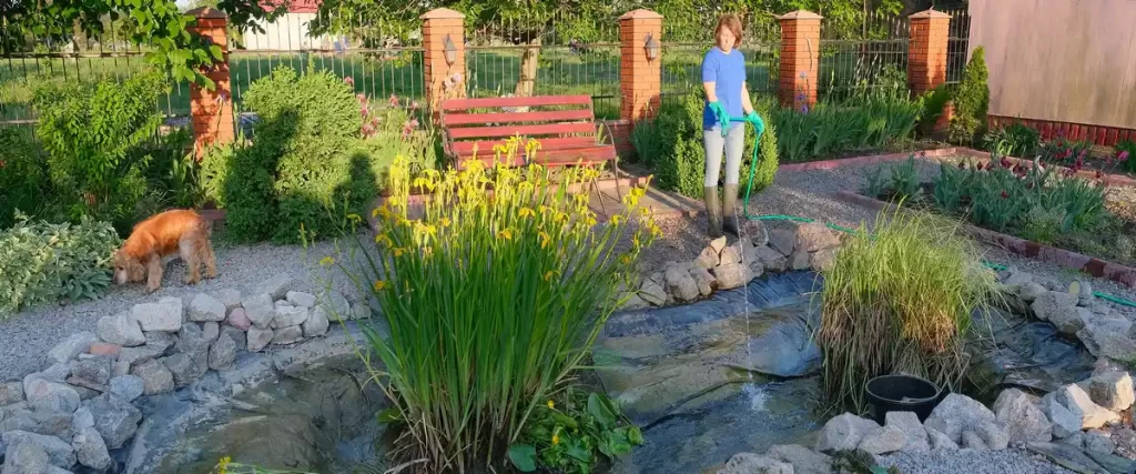 A woman wearing rubber boots and gloves using a garden hose to fill or clean a stone-bordered backyard pond.