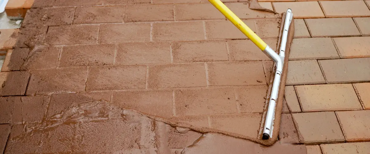 Using a long-handled squeegee to spread a thick brown jointing material or slurry across a brick paver surface to fill and stabilize the joints.